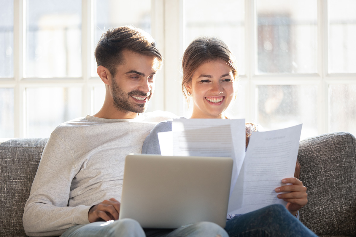 Smiling husband and wife sit on couch using laptop taking care of utility bills and house maintenance documents, happy young couple relax on sofa read paperwork, pay bank credit online via internet