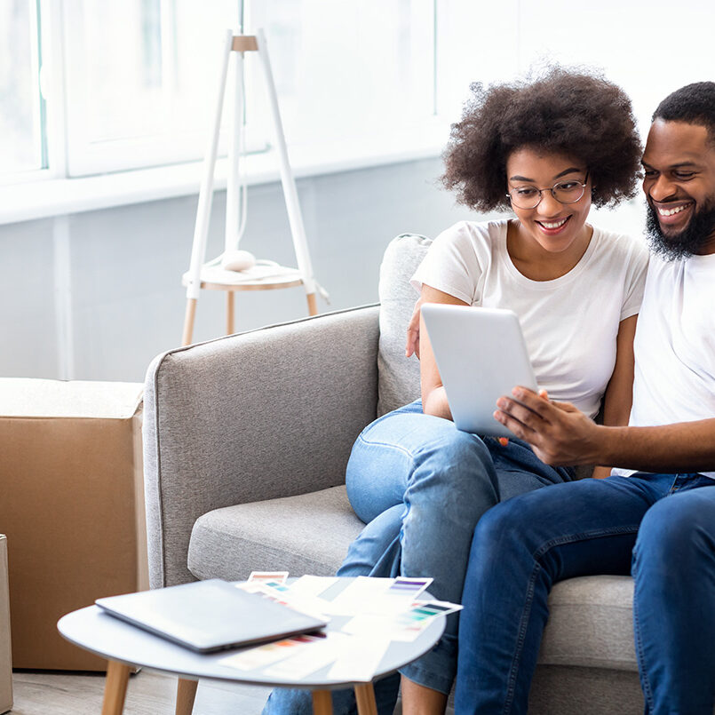 Real Estate Application. Cheerful African Spouses Using Tablet Computer Searching Apartment For Rent Online Sitting Together On Sofa At Home. House Hunting And Relocation Concept